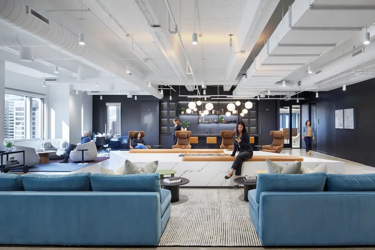 A professional woman sits on a bench near a fire pit in a lounge space in a modern office. 