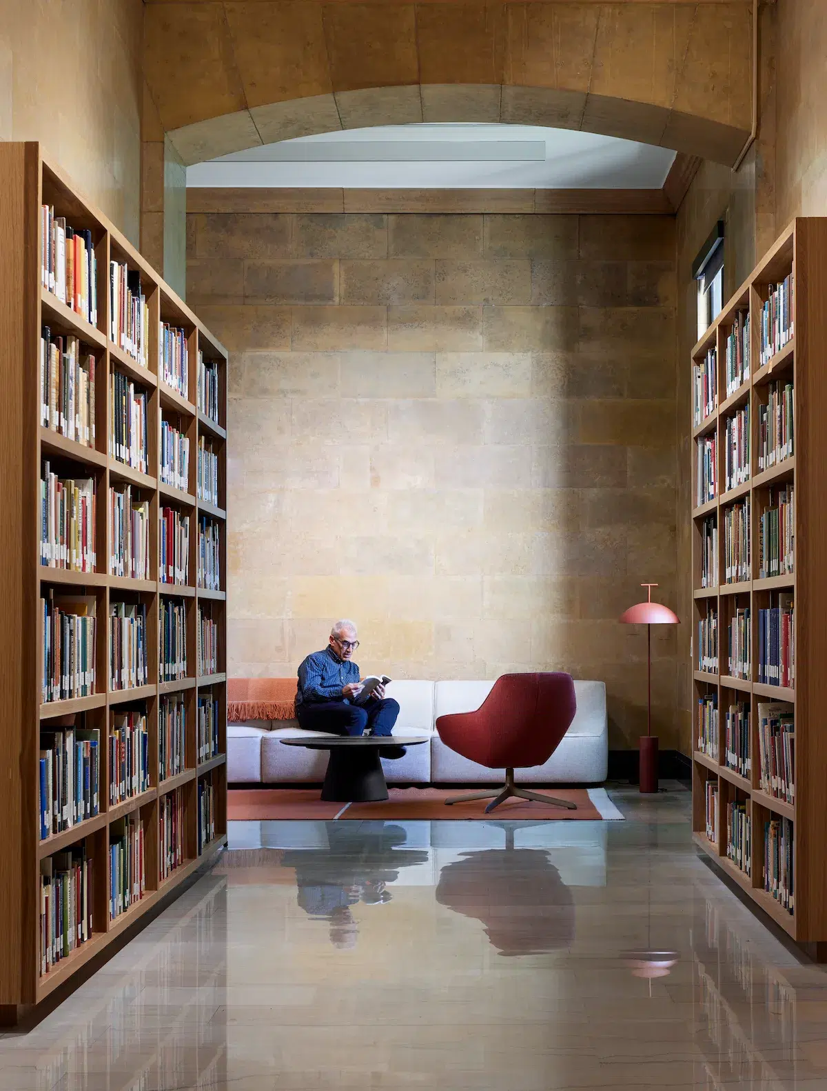 A man sits at the far end of a hall lined with bookshelves in an upscale museum space.