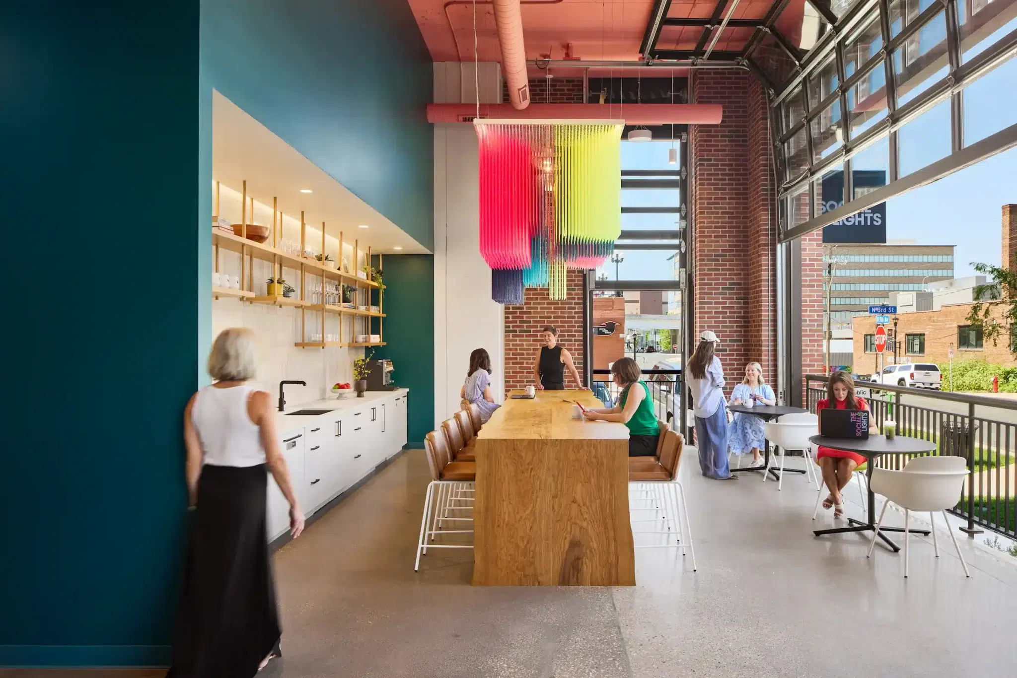 People work in an office cafe space with colorful art/lights and a garage door-style window open to the street. 