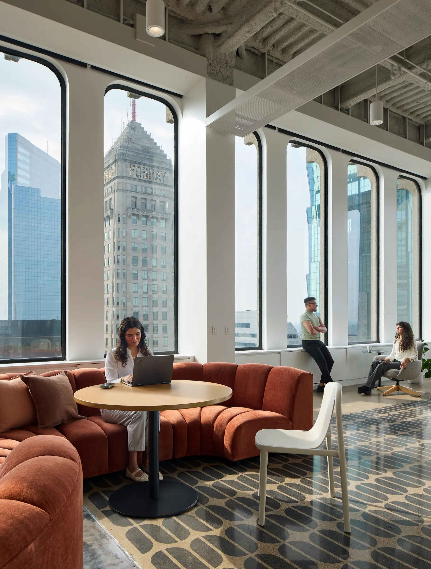 A woman works on a laptop on a red curbed sofa in a modern industrial building with the Foshay building in view.