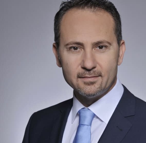 Man in a suit with a blue tie, posing against a plain gray background.