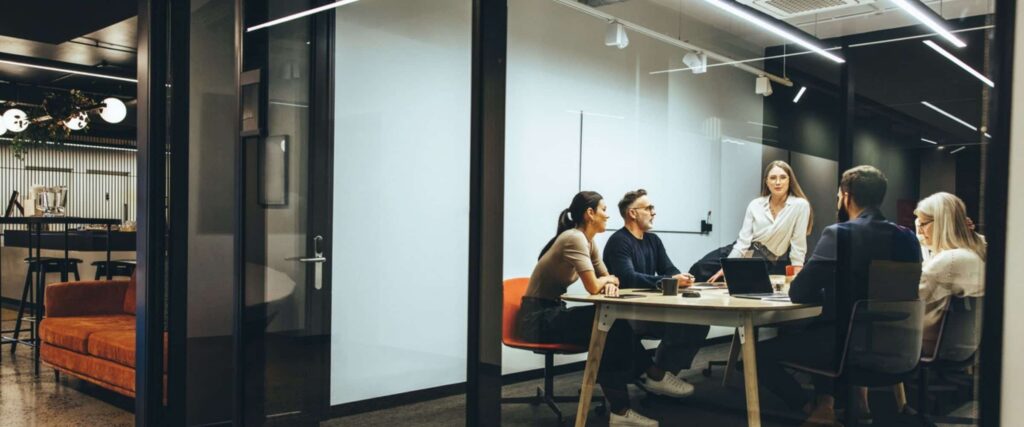 Five people are sitting around a conference table in a modern office, having a meeting. One woman is standing and speaking, while others listen attentively. Laptops and documents are on the table.