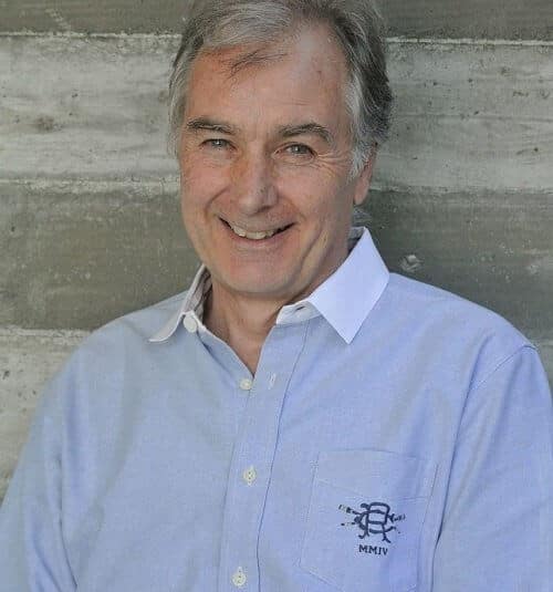 Smiling man with gray hair, wearing a light blue shirt, standing against a gray concrete wall.