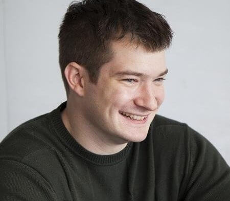 Man with short dark hair, smiling, wearing a dark sweater, against a light background.
