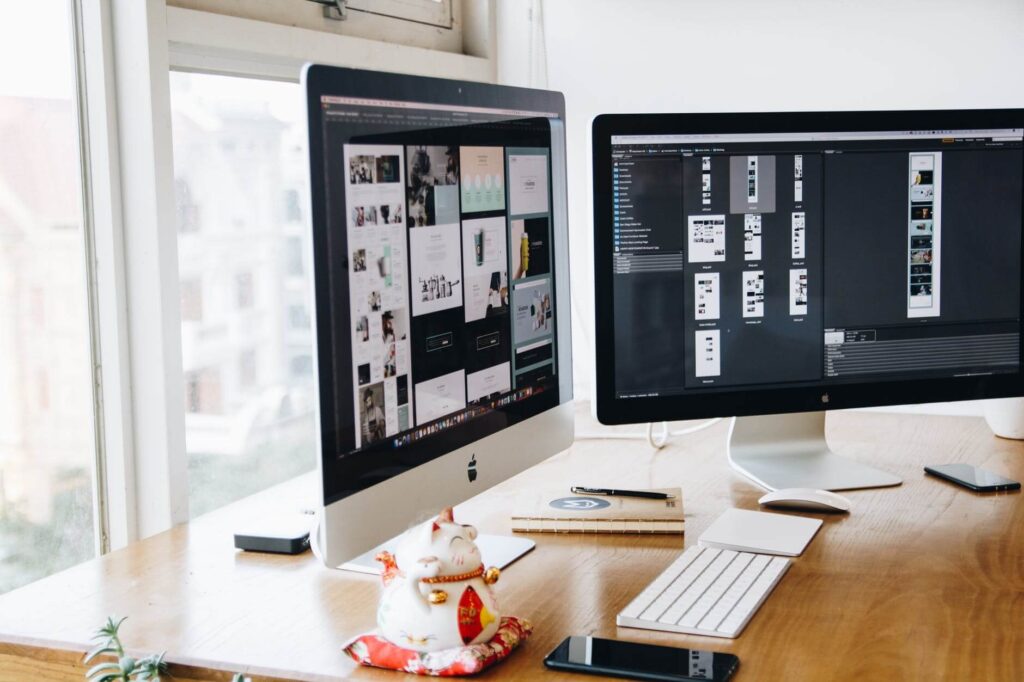 Two desktop monitors displaying design software on a wooden desk with a cat figurine and smartphones nearby.