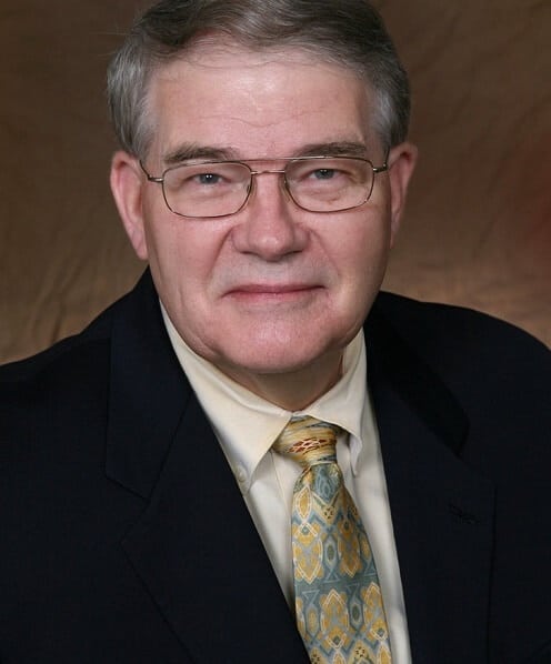 Older male in glasses, wearing a suit and patterned tie, smiles against a brown background.