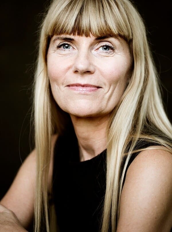 Woman with long blonde hair and bangs, wearing a black top, looking confidently at the camera.