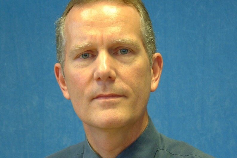 A man in a collared shirt poses in front of a blue background.