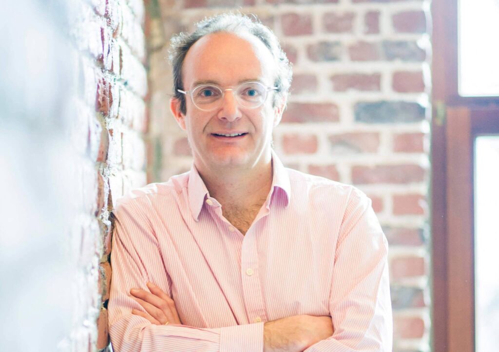 Man in a pink shirt, smiling with arms crossed, leaning against a brick wall near a window.