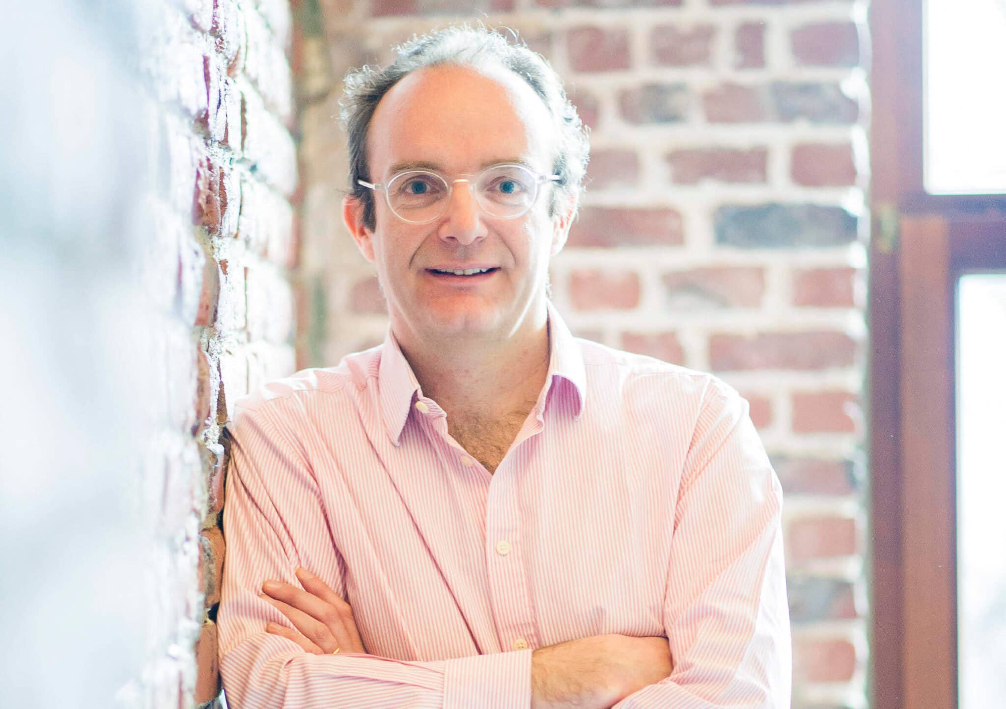 Man in a pink shirt, smiling with arms crossed, leaning against a brick wall near a window.