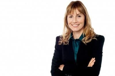 Smiling woman with long hair in a black suit stands with arms crossed against a white background.
