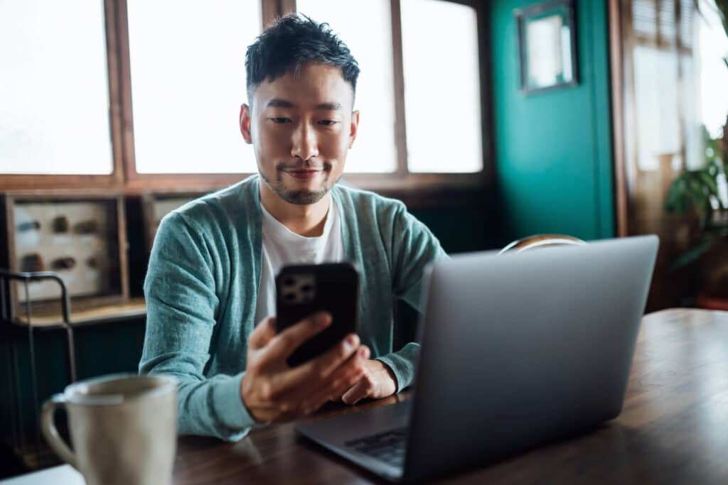 Man sitting at a table with a laptop, looking at his smartphone, in a cozy room with green walls and wooden windows.