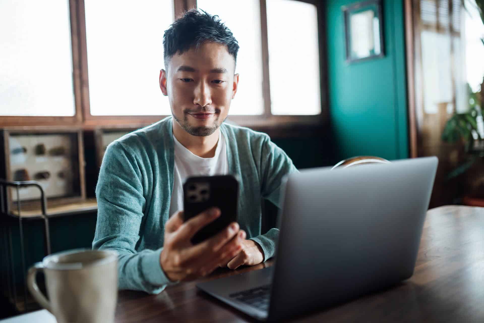 Man sitting at a table with a laptop, looking at his smartphone, in a cozy room with green walls and wooden windows.