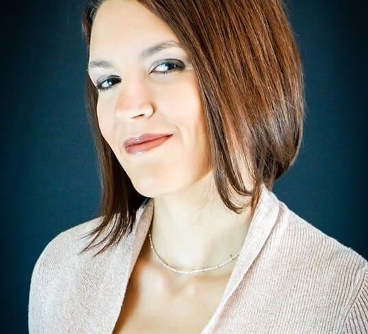 Woman with brown hair, wearing a light sweater and necklace, smiling slightly against a dark background.