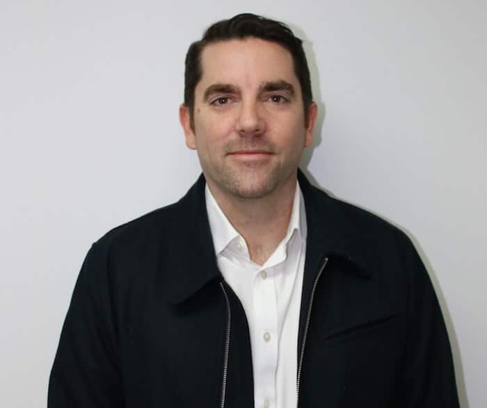 Man in a white shirt and dark jacket, standing against a plain white background.