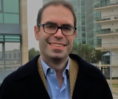 A person wearing glasses, smiling outdoors on a foggy day, with modern buildings in the background.