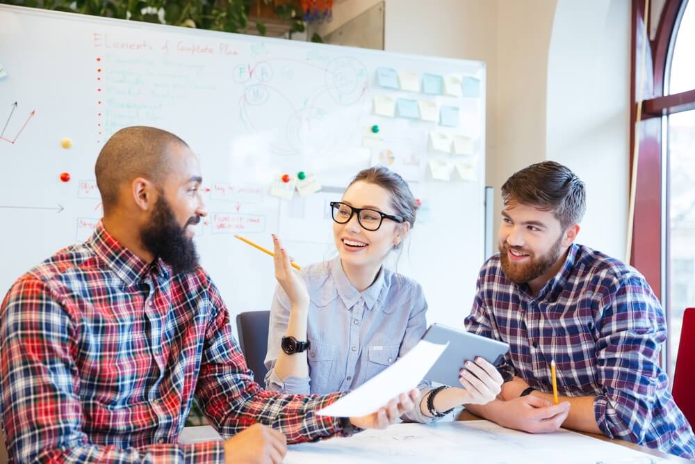 Three people smiling and talking at a table with papers. A whiteboard with notes is in the background.