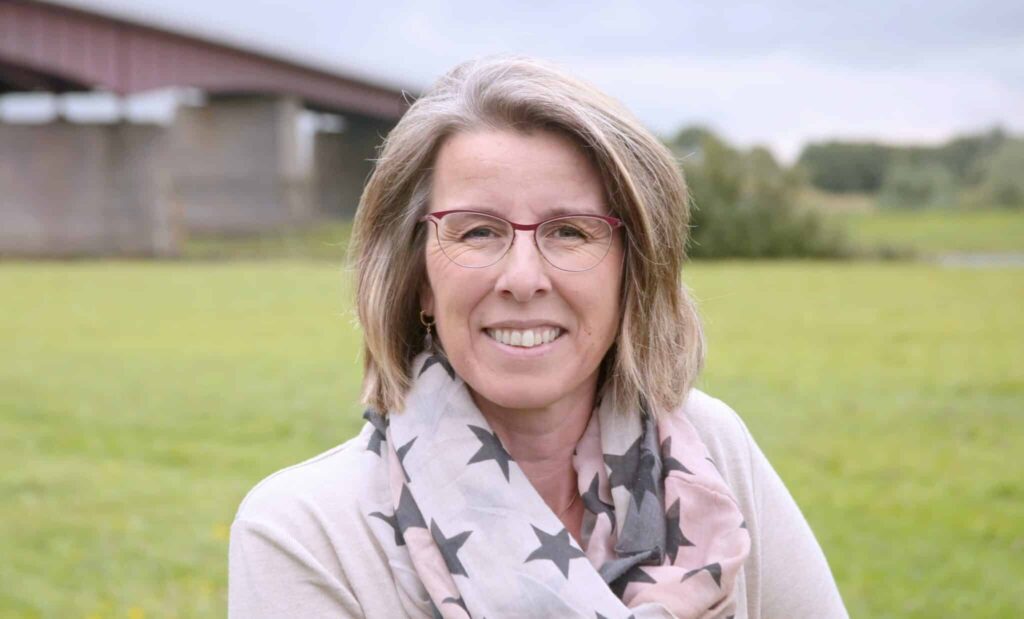 Smiling woman with glasses and star-patterned scarf stands outdoors, with a grassy field and bridge in the background.