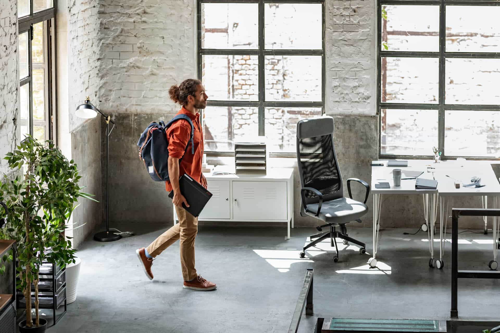 A person with a backpack and laptop walks into a modern office with large windows and a desk.