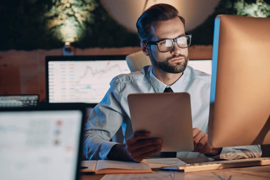 A man in glasses works at a desk with computers, holding a tablet, in a dimly lit office.
