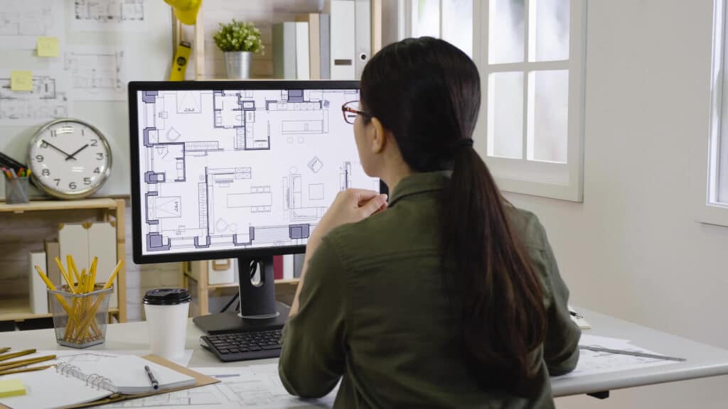 A person with long hair works at a desk, examining architectural plans on a computer monitor in an office setting.