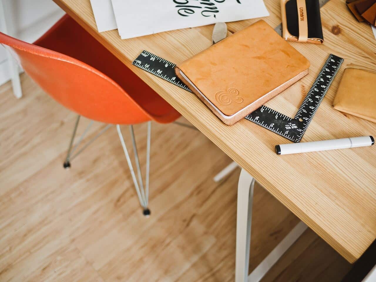A wooden desk with a leather notebook, rulers, a marker, and an orange chair on a wooden floor.
