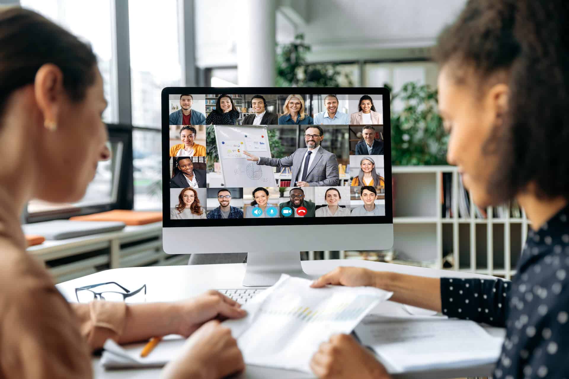 Two people in a video conference, looking at documents. A virtual meeting with many participants is on the computer screen.