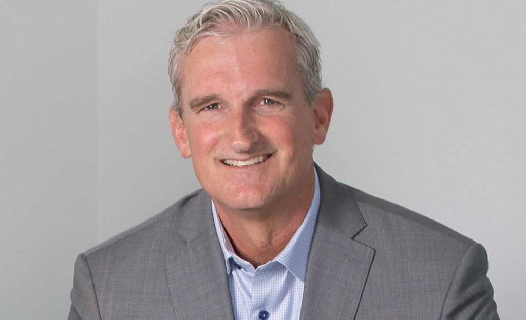 Smiling man with gray hair in a gray suit and blue shirt against a plain background.
