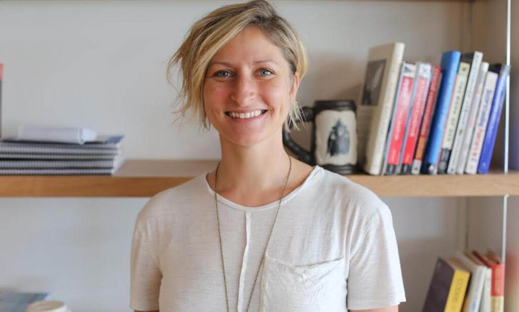 Person smiling, wearing a white shirt, with short hair. Standing in front of a bookshelf with various books and items.