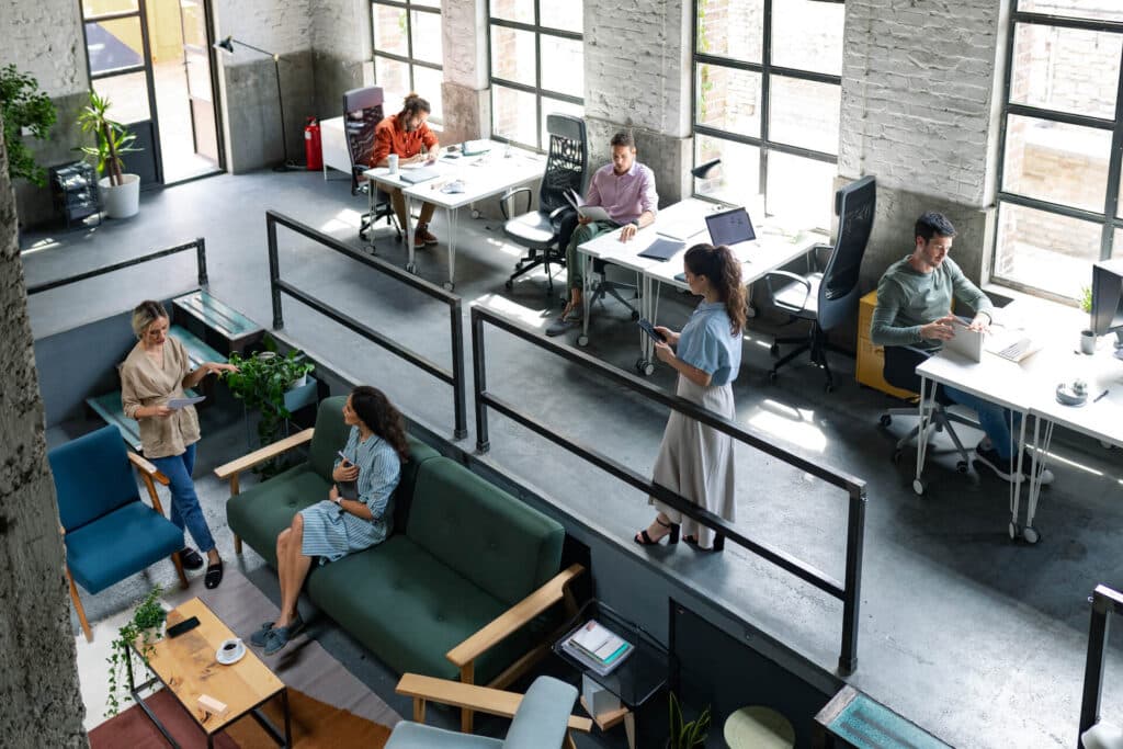 Open office space with people working at desks and sitting on couches, with large windows and plants around.