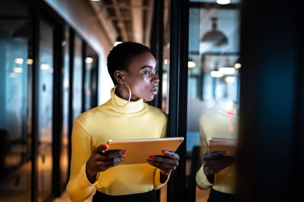 Person in a yellow sweater holding a tablet, standing in a corridor with glass walls, looking thoughtfully to the side.