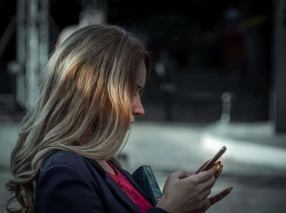 Woman with long hair, in a dark coat, looking at her phone in an outdoor setting.