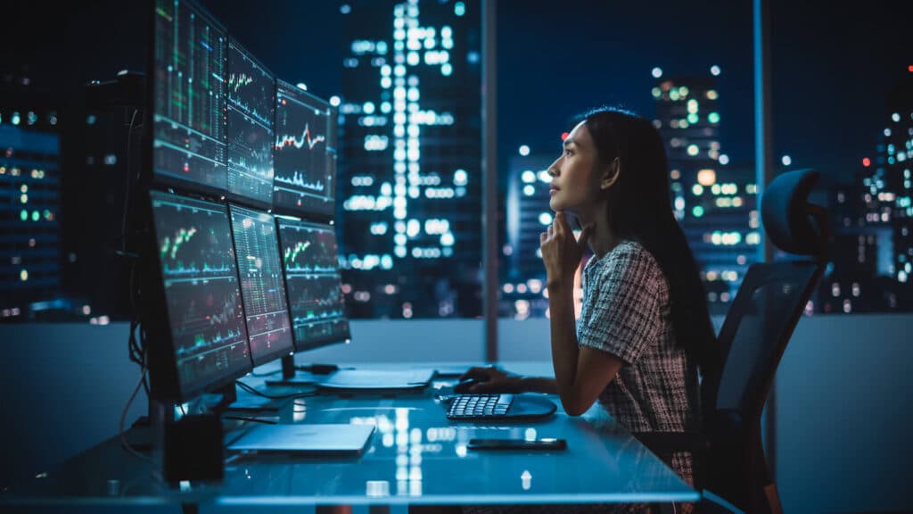Person analyzing financial charts on multiple monitors in an office at night, city lights visible through windows.