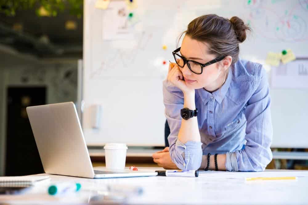 A woman in glasses leans over a table, looking at a laptop. A coffee cup is nearby, and a whiteboard is in the background.