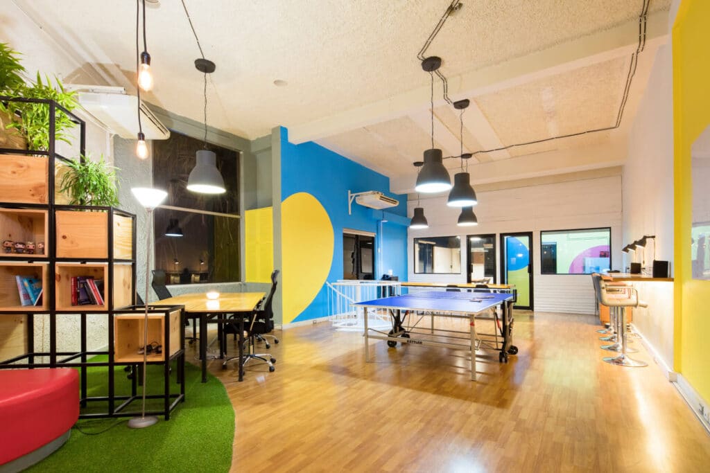 Modern office space with a ping pong table, vibrant blue and yellow walls, and a seating area with bookshelves.