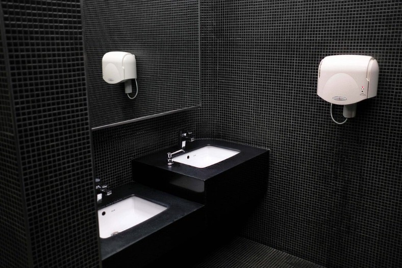 Modern public restroom with black tiled walls, two white sinks with faucets, a large mirror, and a white automatic hand dryer mounted on the wall—an example of universal design in the workplace that ensures accessibility and comfort for everyone.