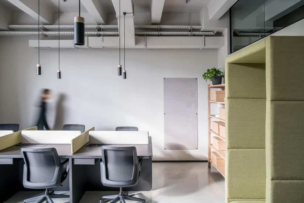 Modern office with empty desks, swivel chairs, pendant lights, a blurred figure walking, and a green partitioned space.