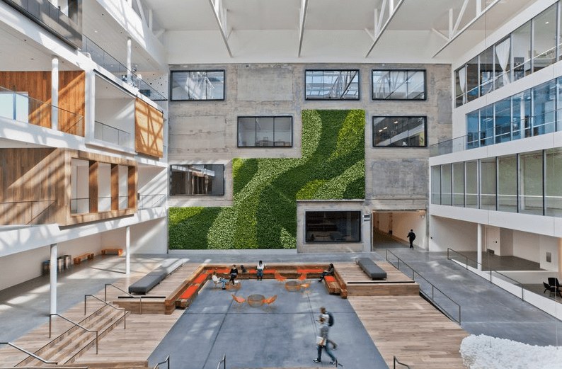 A modern atrium features a green living wall, large windows, wooden accents, tiered seating, and open spaces with people walking and sitting—showcasing the inspiration behind contemporary design office space. Natural light streams through the glass ceiling.