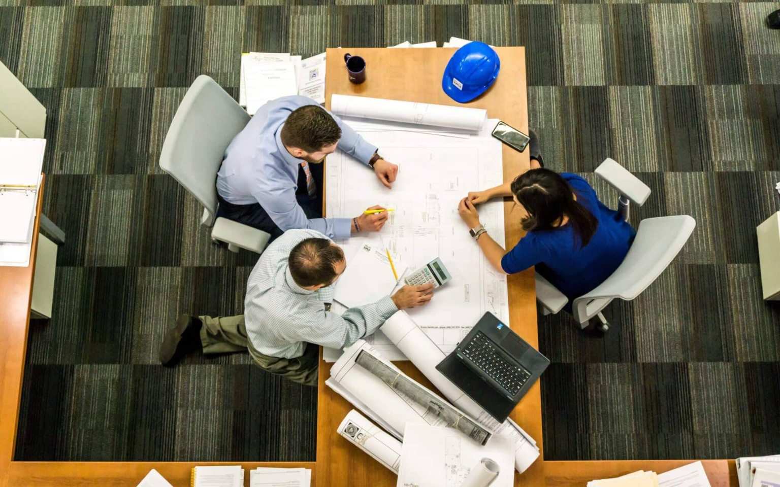 Three people sit at a table reviewing architectural plans, collaborating on office facility management strategies. Blueprints, a blue hard hat, laptop, phone, calculator, and coffee mug are on the table. The scene is viewed from above in an office setting.