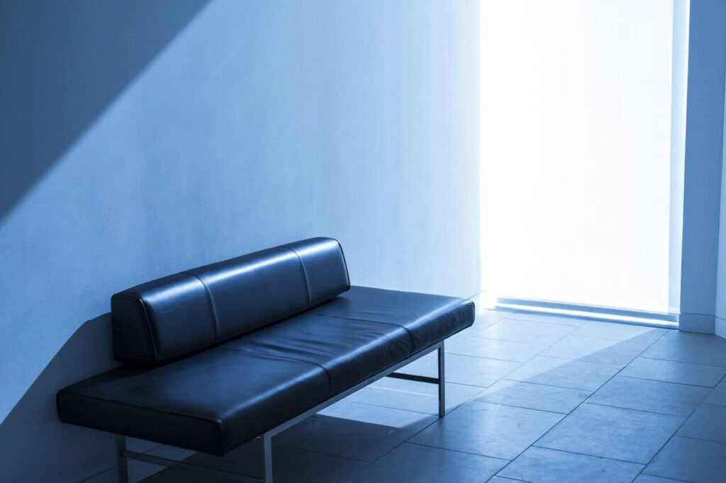 A minimalist black leather bench sits in a mostly empty, sunlit room with blue walls and tiled floor. Light streams in from a large window, casting shadows on the bench and wall.