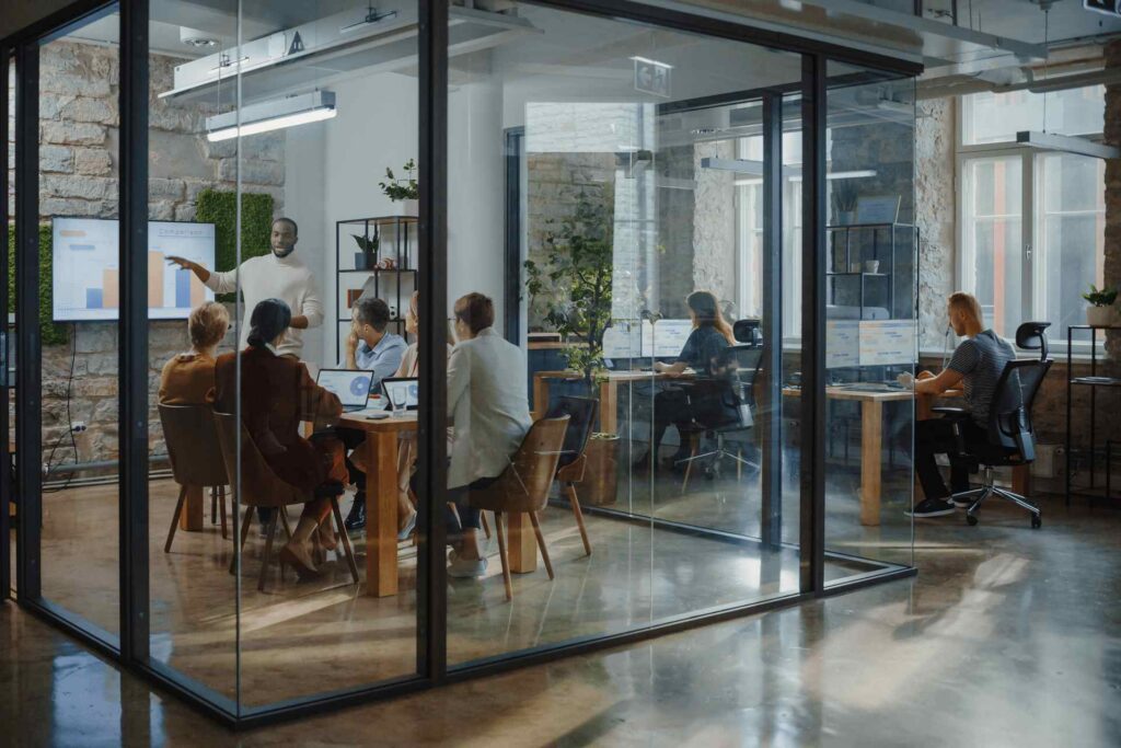A group of people sit at tables in a glass-walled office, listening to a person give a presentation with a chart on a screen focused on space utilization. Others work on computers in the background.
