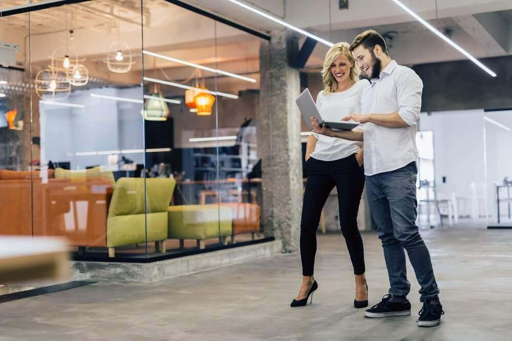 Two people smiling and looking at a laptop in a modern office with colorful furniture behind them.