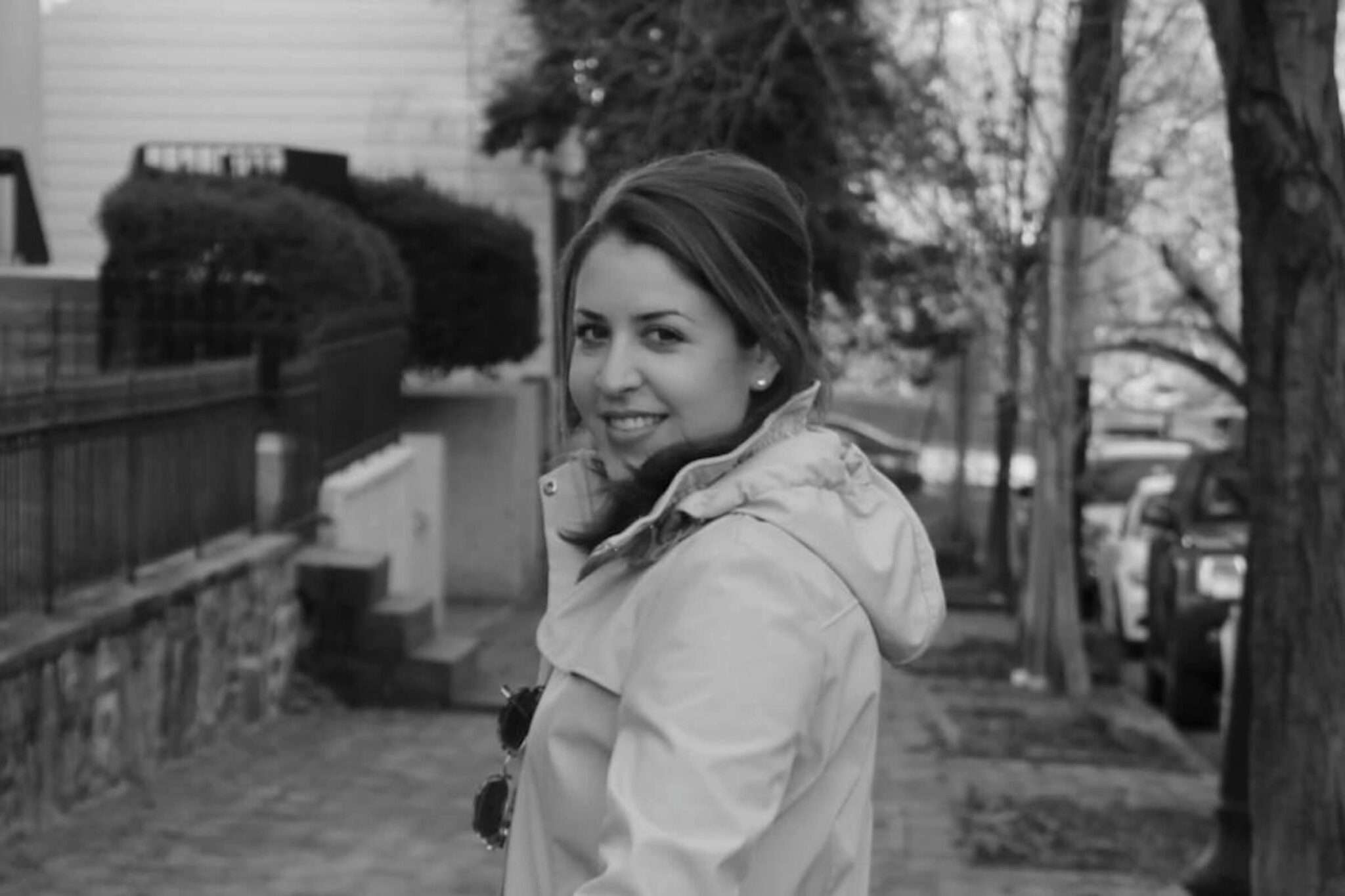A woman in a light coat looks back and smiles while walking on a sidewalk lined with trees and parked cars in a residential neighborhood. The photo is in black and white.