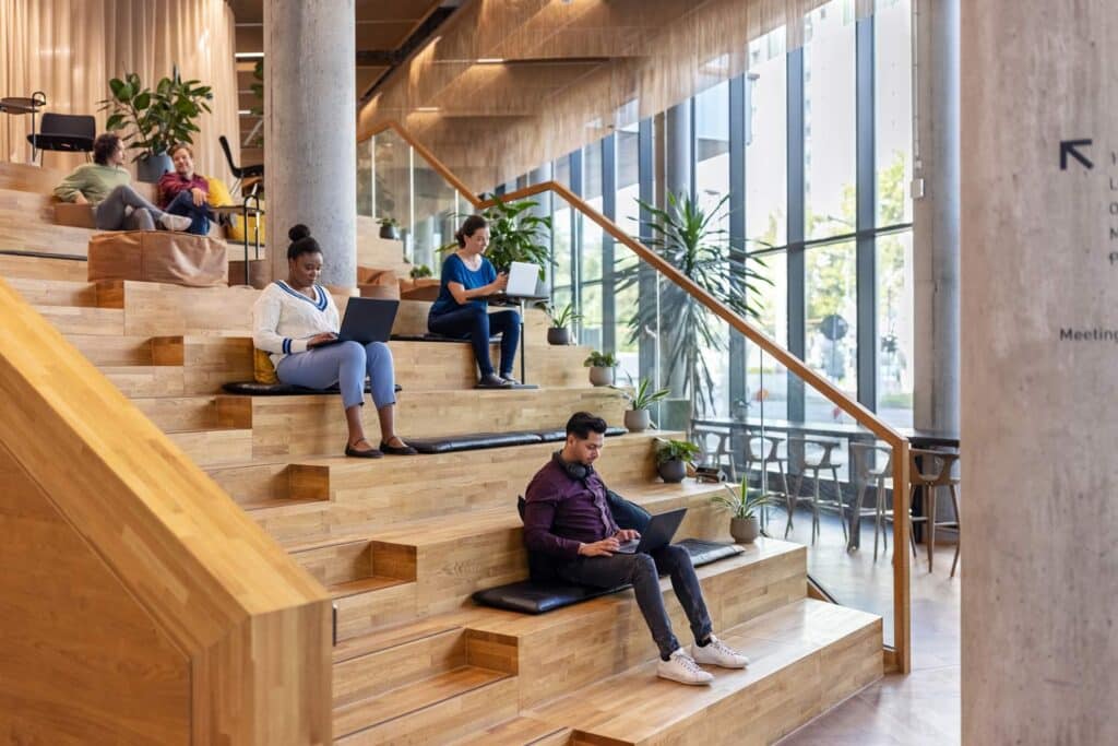People working on laptops, seated on wooden steps with cushions in a modern, open space with large windows and plants.