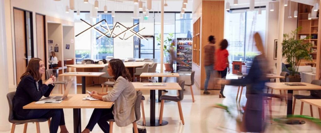 Modern office with people sitting at tables, having discussions—some even engaged in hybrid workplace platform comparison as part of their collaboration. Others walk or ride a bike through the space. Large windows, wooden accents, and shelves create a bright, open, and collaborative atmosphere.