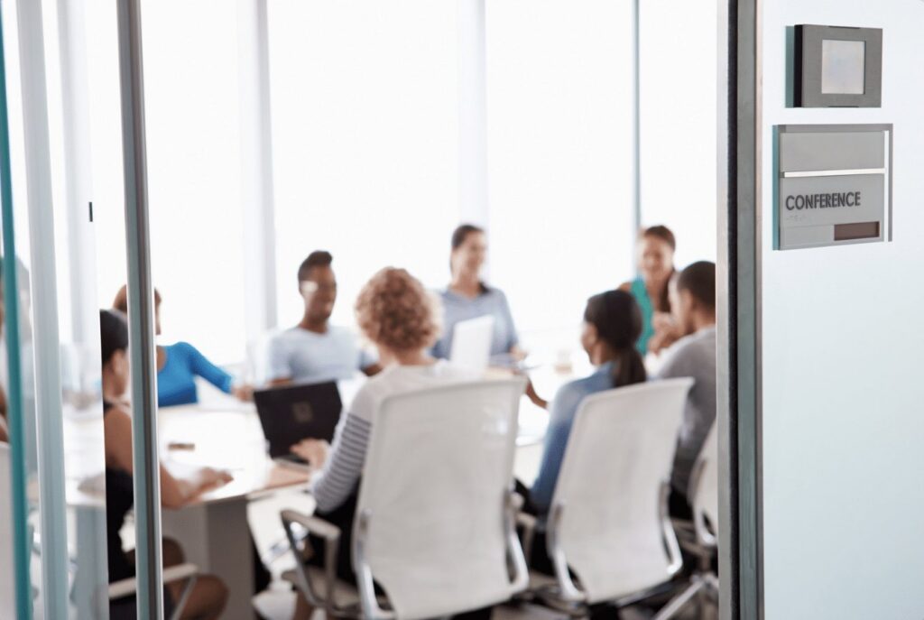 People having a meeting in a conference room with a sign on the door visible in the foreground.
