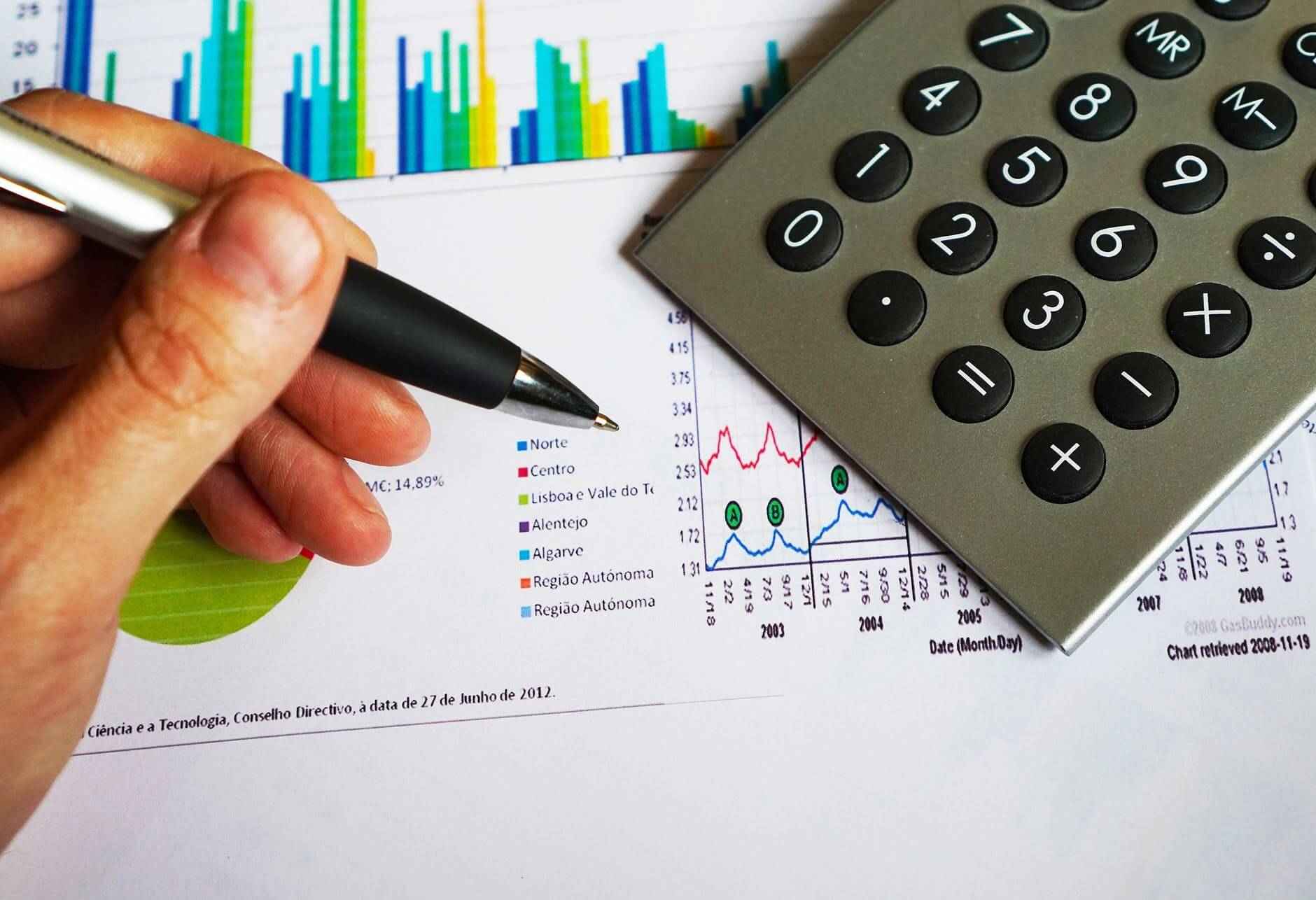 A hand holding a pen points at a financial chart, with a calculator resting on printed graphs featuring colorful bars and lines.