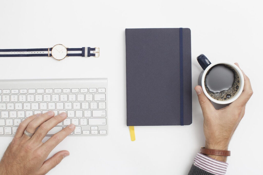 Hands typing on a keyboard, holding a coffee mug; watch and notebook are on the table.