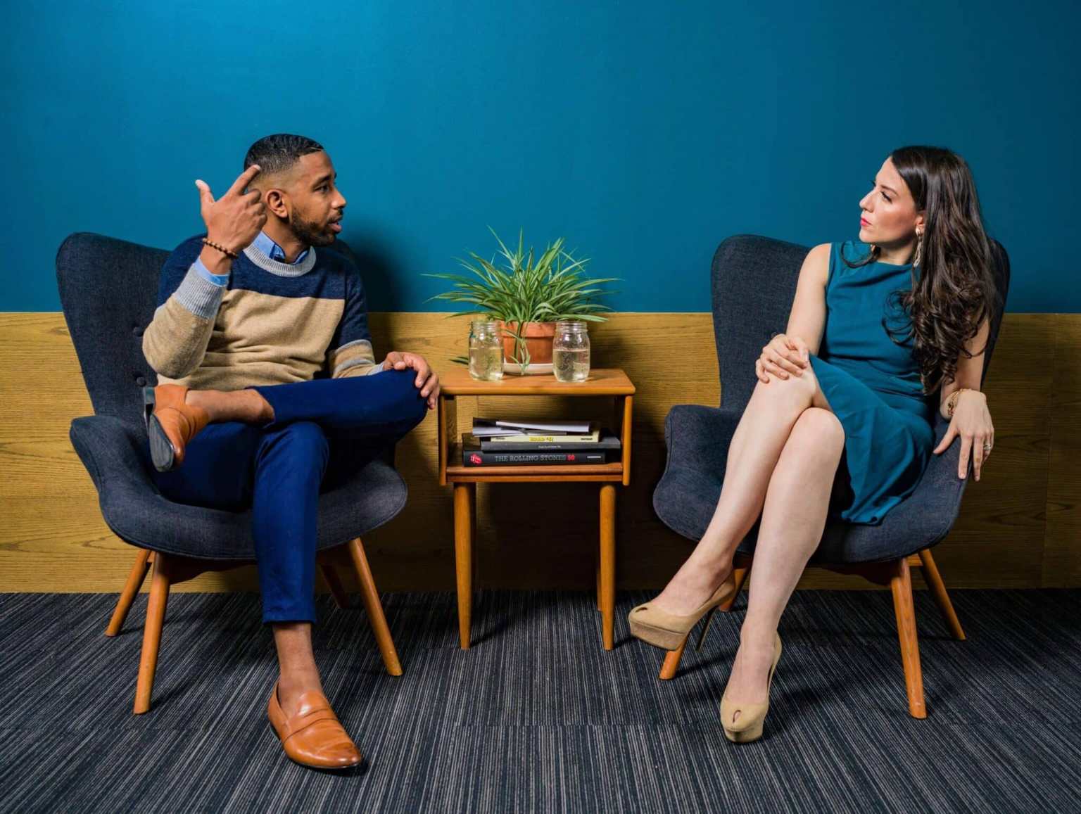A man and a woman sit in armchairs, facing each other and talking. A small table with books, two glasses, and a plant is between them. The setting appears casual and office-like, with a blue and wood wall behind them.