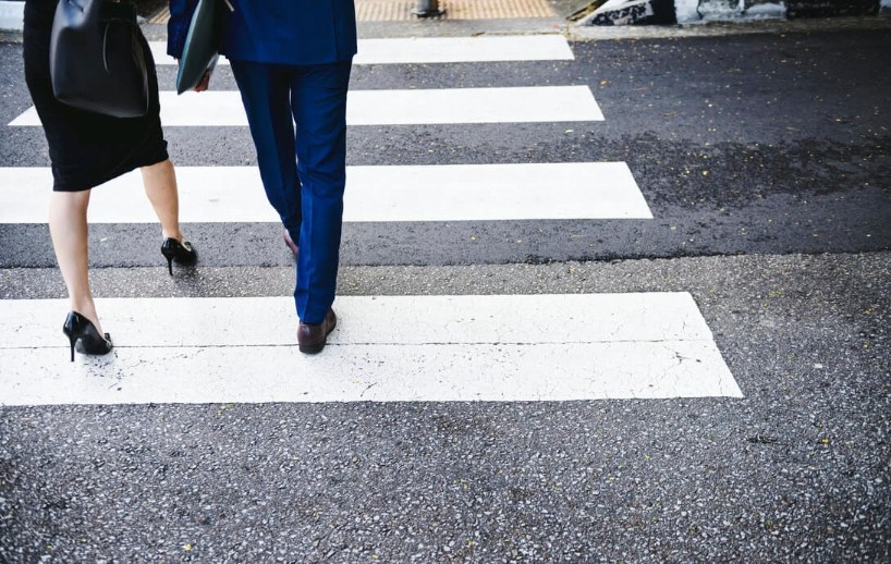Two people in business attire walk across a crosswalk, photographed from behind at street level. The pavement and white stripes of the crosswalk are visible.